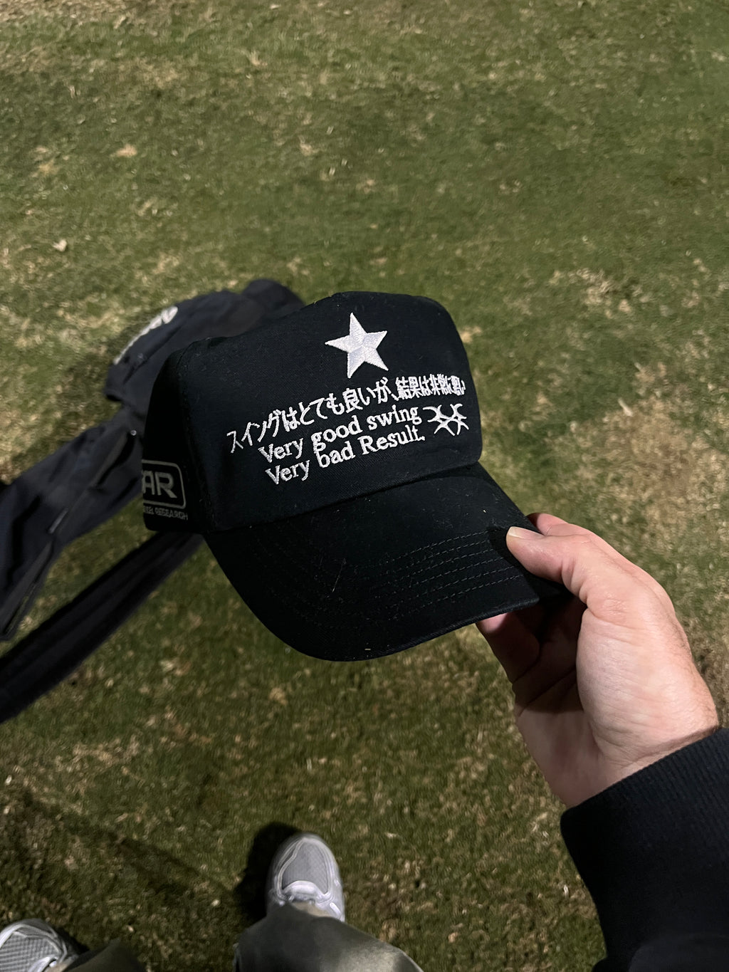 Black golf cap with white text and star logo held by a hand on a grassy golf course